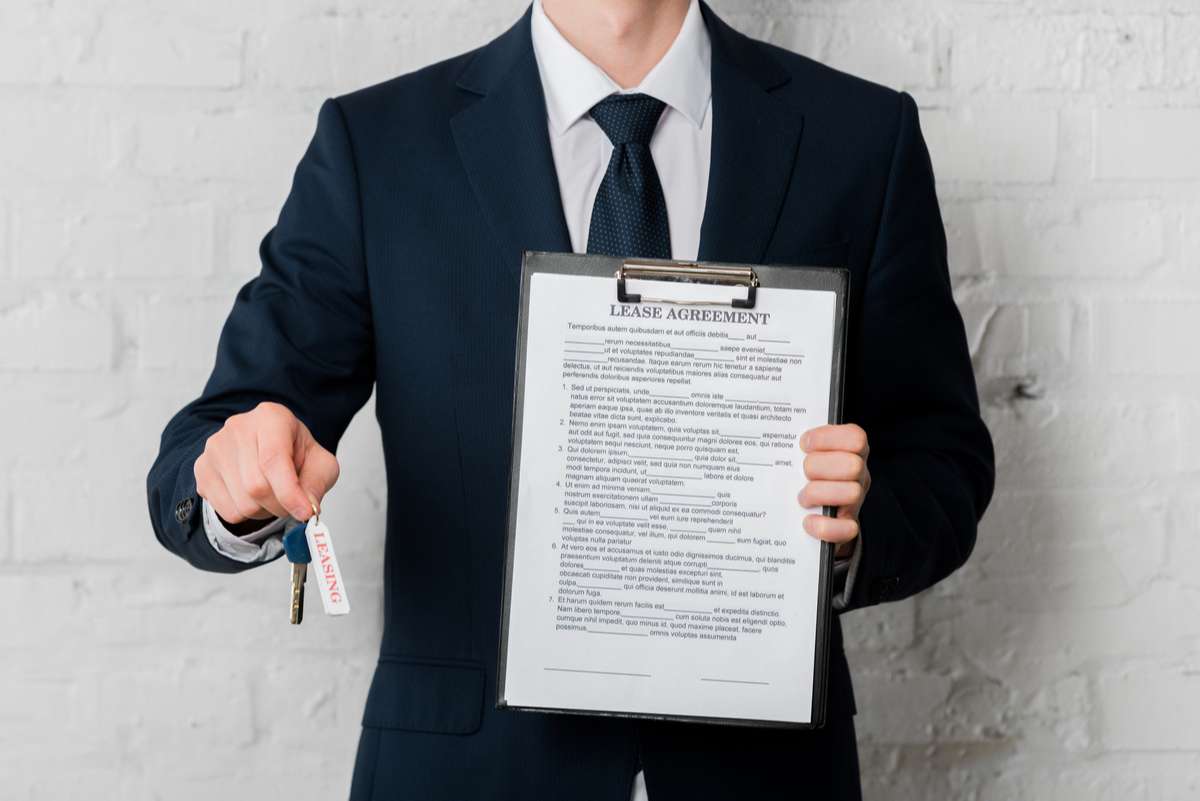 Cropped view of realtor in suit holding key with leasing lettering and clipboard with lease agreement on white