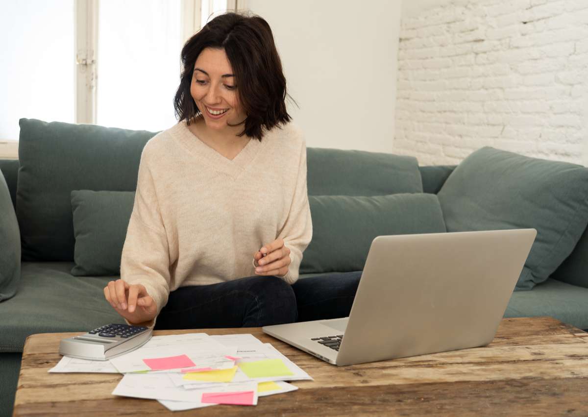 Happy young woman sitting on sofa surrounded by papers calculating expenses and paying bills