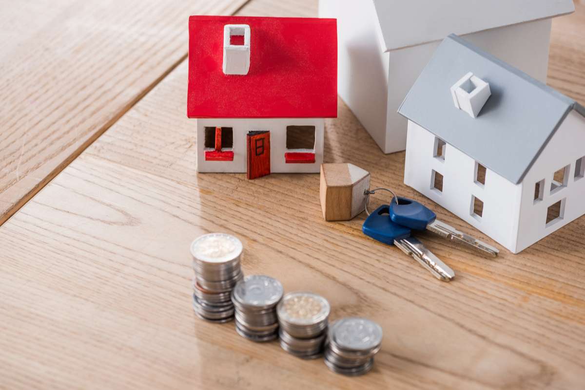 House models near keys and stacks of golden and silver coins on wooden table