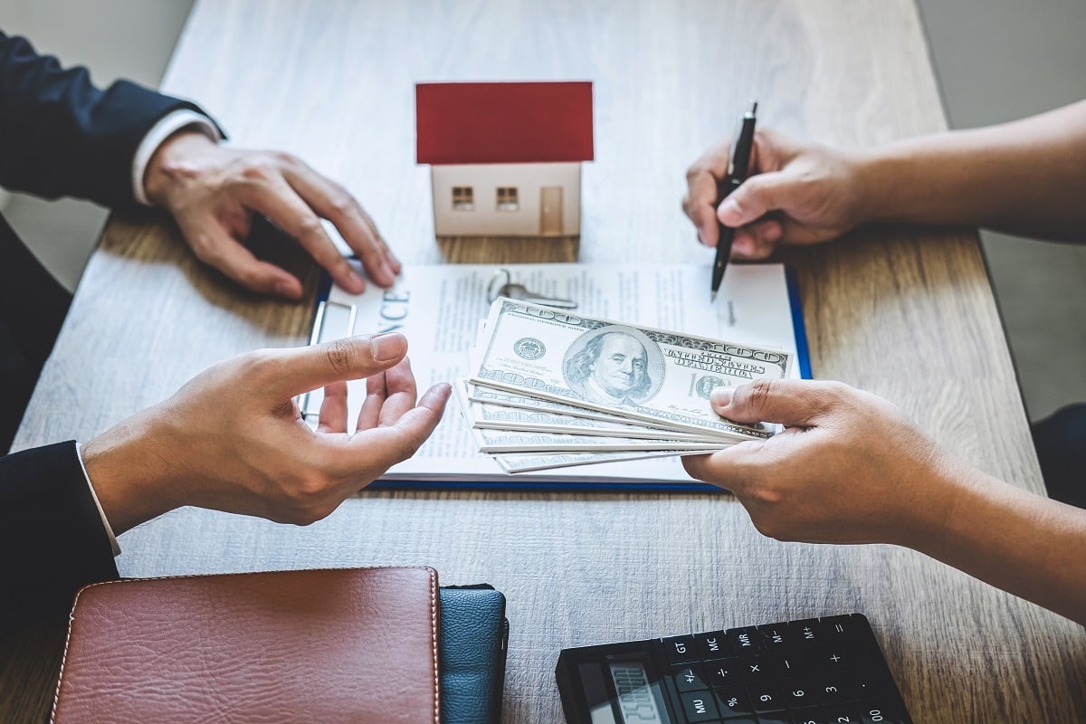 Someone handing money to a person and signing a contract next to a small house and a calculator