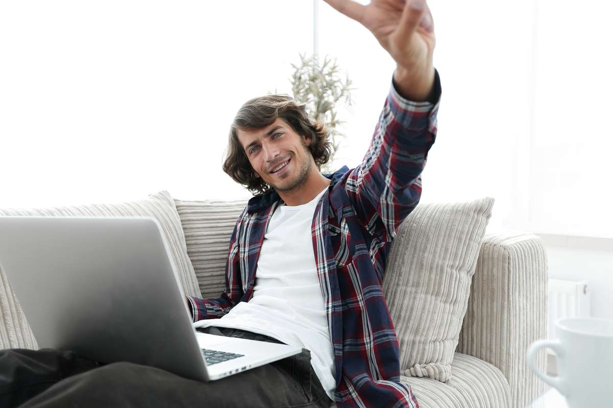 Happy guy with laptop sitting on sofa and showing his hand a winning gesture