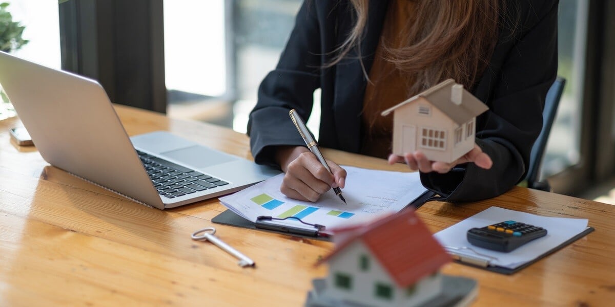 A professional woman writing and holding a small house next to a laptop