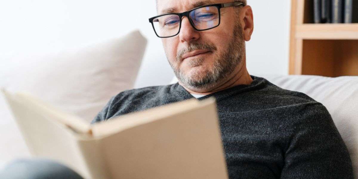 Pleased adult unshaven man in eyeglasses reading book while resting at home